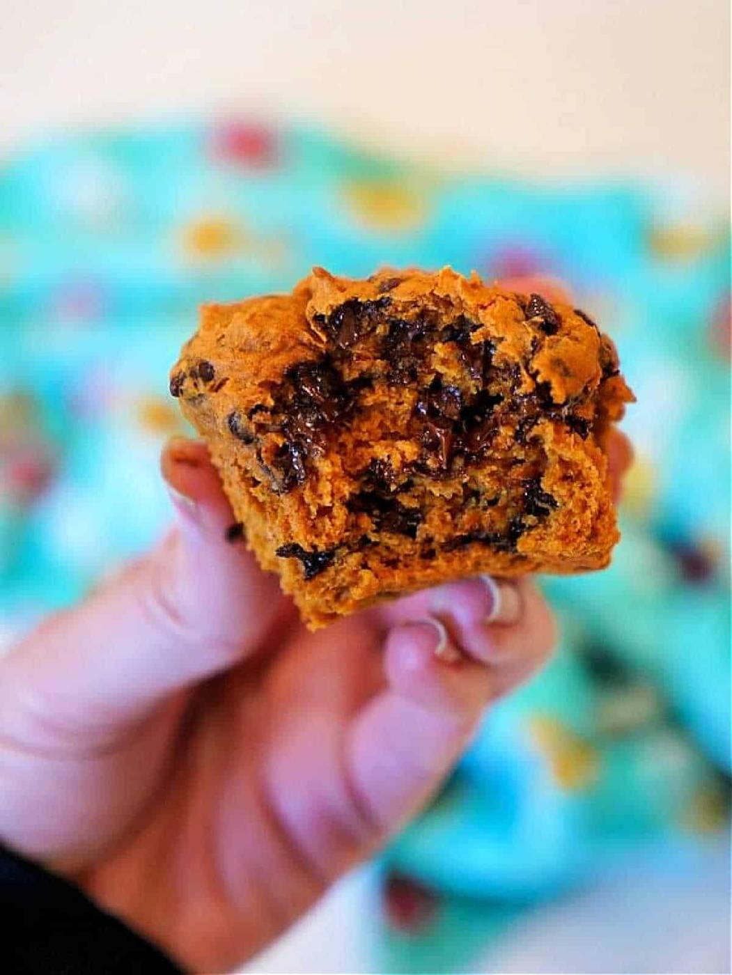 A hand holds a pumpkin chocolate chip muffin with a bite taken out, its rich flavors reminiscent of 2 ingredient pumpkin muffins, set against a blurry, colorful background.