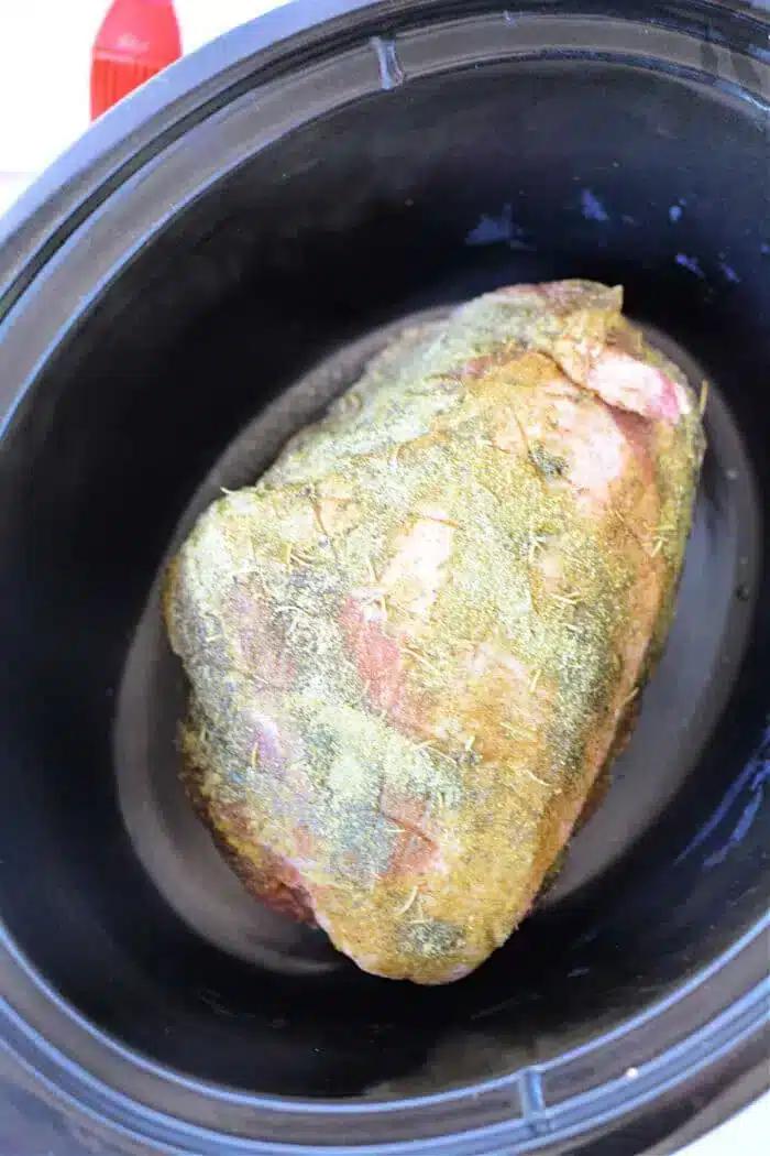 A seasoned piece of pork is placed inside a slow cooker. The 3 Ingredient Pork Roast is covered in herbs and spices, ready to be cooked.