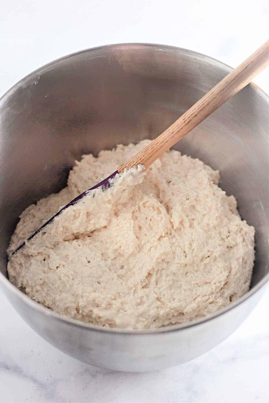 A mixing bowl filled with bread dough and a wooden spoon set beside it on a pristine white surface fuses simplicity and charm, reminiscent of crafting 4 Ingredients Basic Biscuits from scratch.