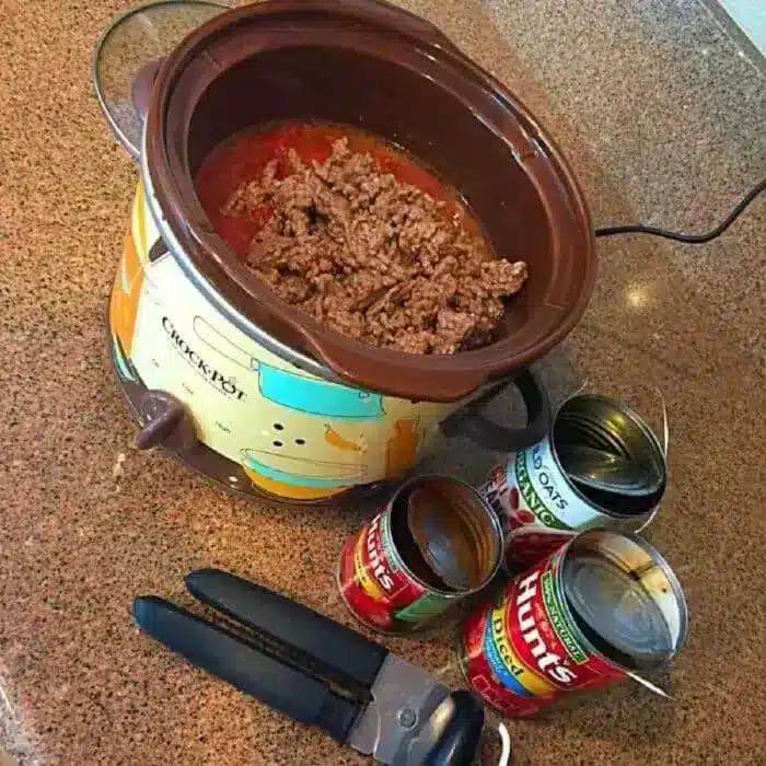 A slow cooker with ground beef inside is on a countertop. Next to it are three opened cans of Hunts tomato sauce and a can opener, hinting at an easy 5 ingredient crockpot chili recipe.