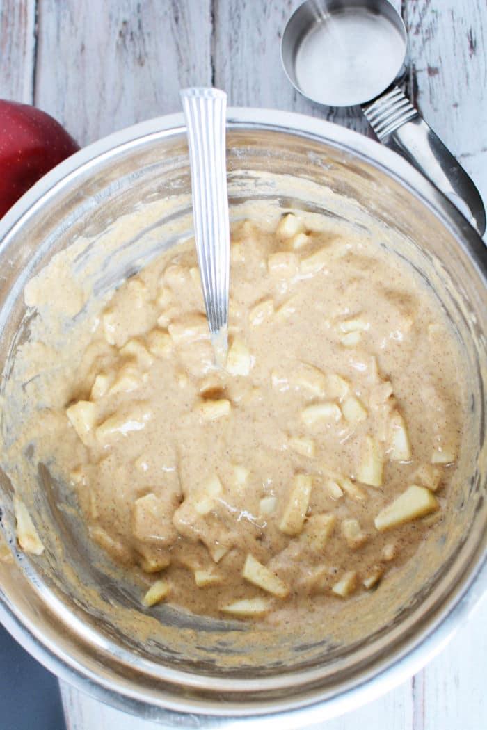 Bowl of apple fritter batter with a spoon, ready to transform into air fryer apple fritters, beside a measuring cup and red apple on a wooden surface.