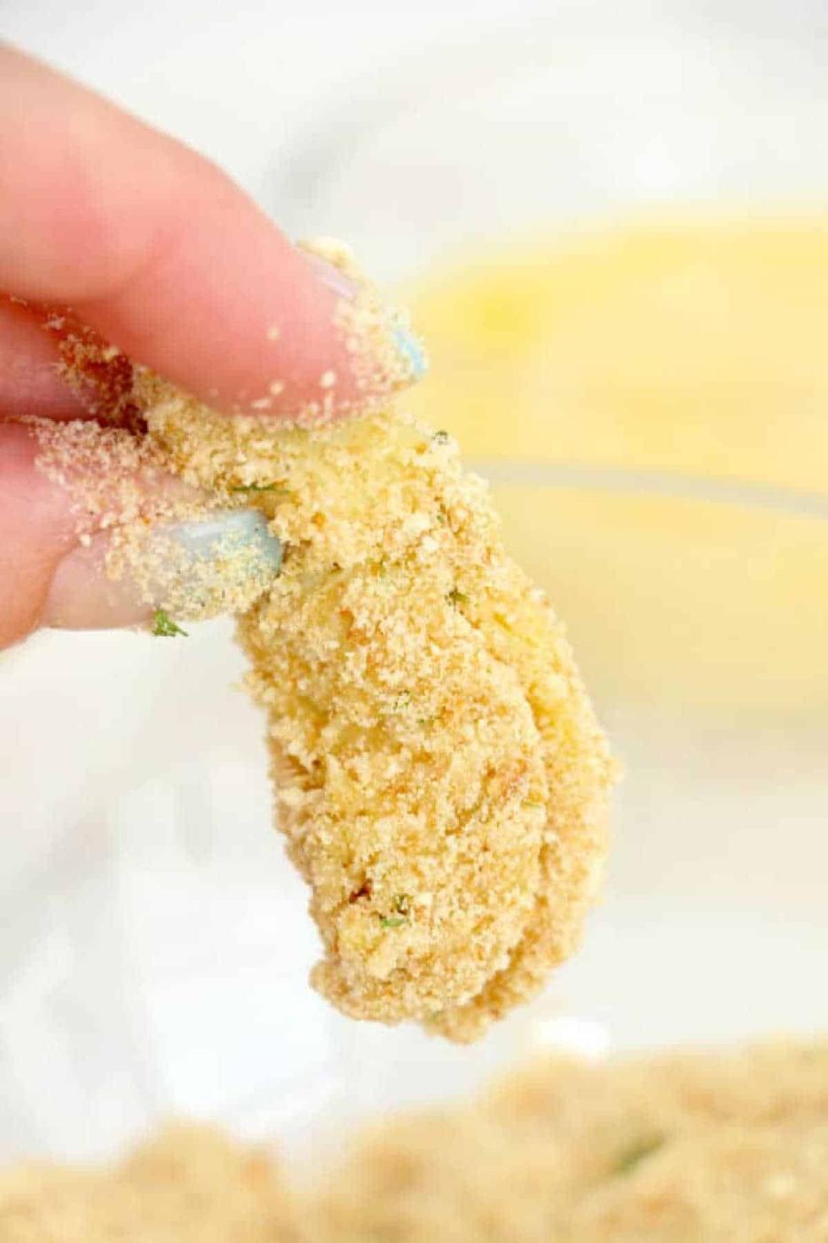 A hand holding a breaded, uncooked slice of zucchini above a bowl of breadcrumbs, ready to be cooked alongside crispy air fryer artichoke hearts.