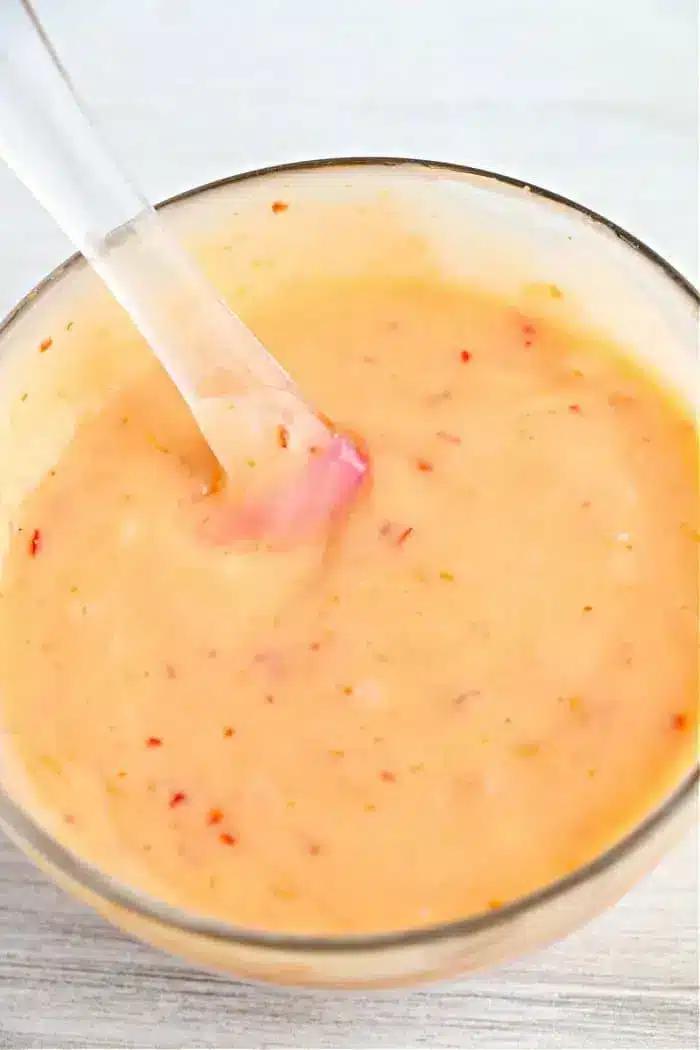A clear bowl filled with a creamy bang bang sauce containing red flecks, reminiscent of the perfect dip for air fryer bang bang shrimp. A transparent spoon is partially submerged in the sauce, and the background is a white surface.