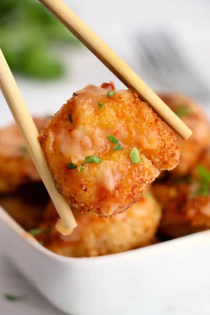 A close-up of chopsticks holding a piece of breaded and fried shrimp, garnished with chopped herbs and a light sauce that gives it an almost bang bang shrimp vibe. In the background, there is a square white bowl filled with more air-fried pieces of the same delicious breaded tofu.