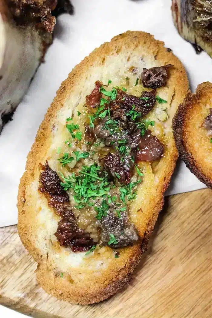 A slice of toasted bread topped with chopped beef and rich bone marrow, garnished with finely chopped parsley, displayed on a wooden surface.
