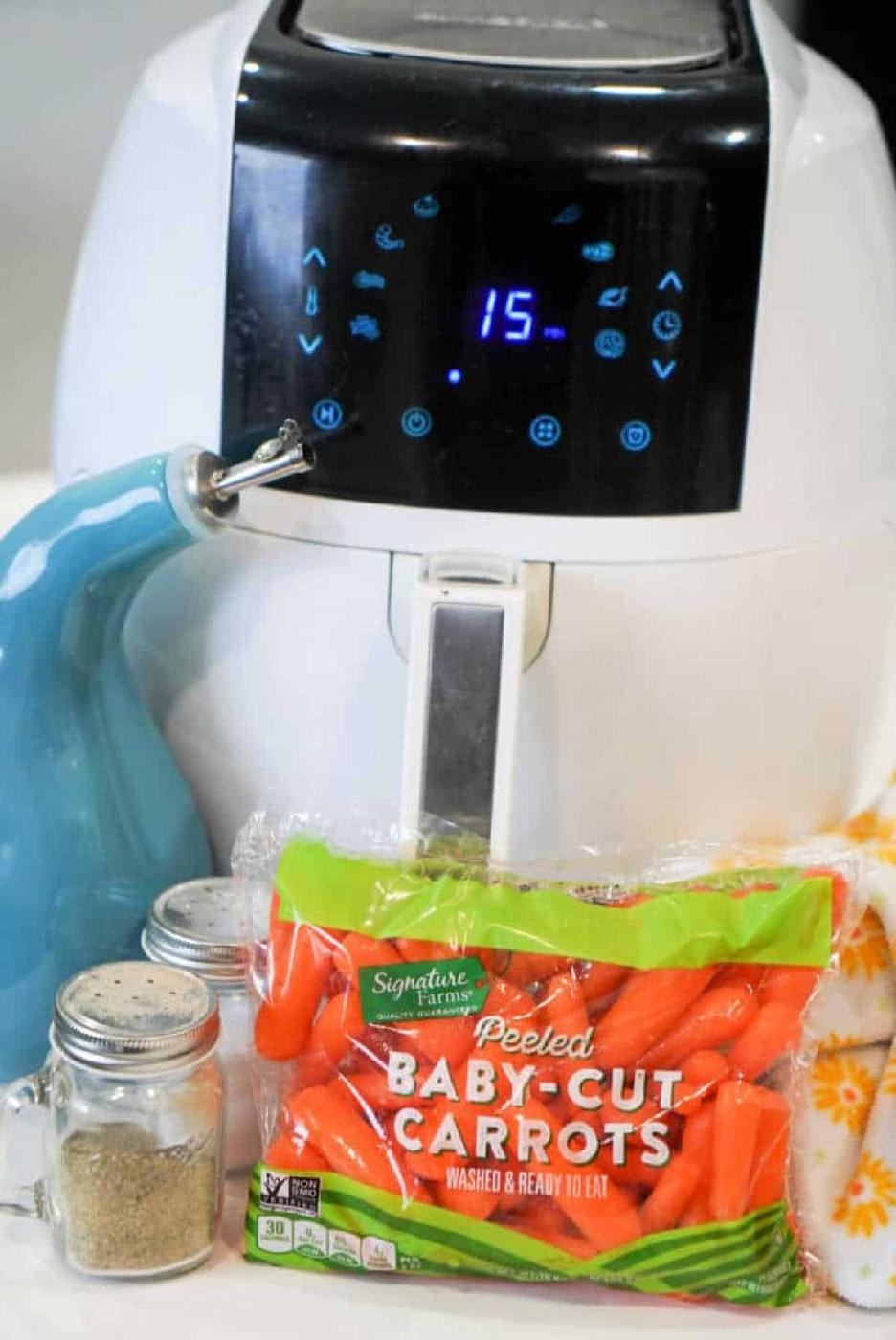 Bag of baby carrots, seasonings, and oil sit in front of a digital air fryer set to 15 minutes—everything you need for quick and tasty air fryer carrots.