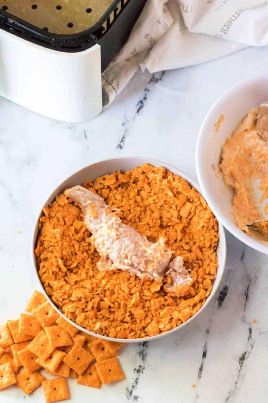 Air Fryer Cheese It Chicken Tenders: Uncooked chicken tender coated in crushed cheese crackers, set beside an air fryer and scattered crackers.