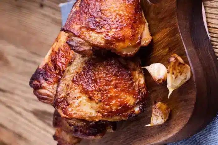 air fried bone-in chicken thighs with crispy, browned skin are arranged on a wooden cutting board. Garlic cloves, also roasted, are placed next to the chicken pieces. The background features a wooden surface with a rustic appearance.