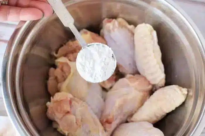 A hand holding a tablespoon of white powder, likely baking powder, hovers over a metal bowl filled with raw chicken wings, ready to be tossed and cooked into crispy air fryer chicken wings.