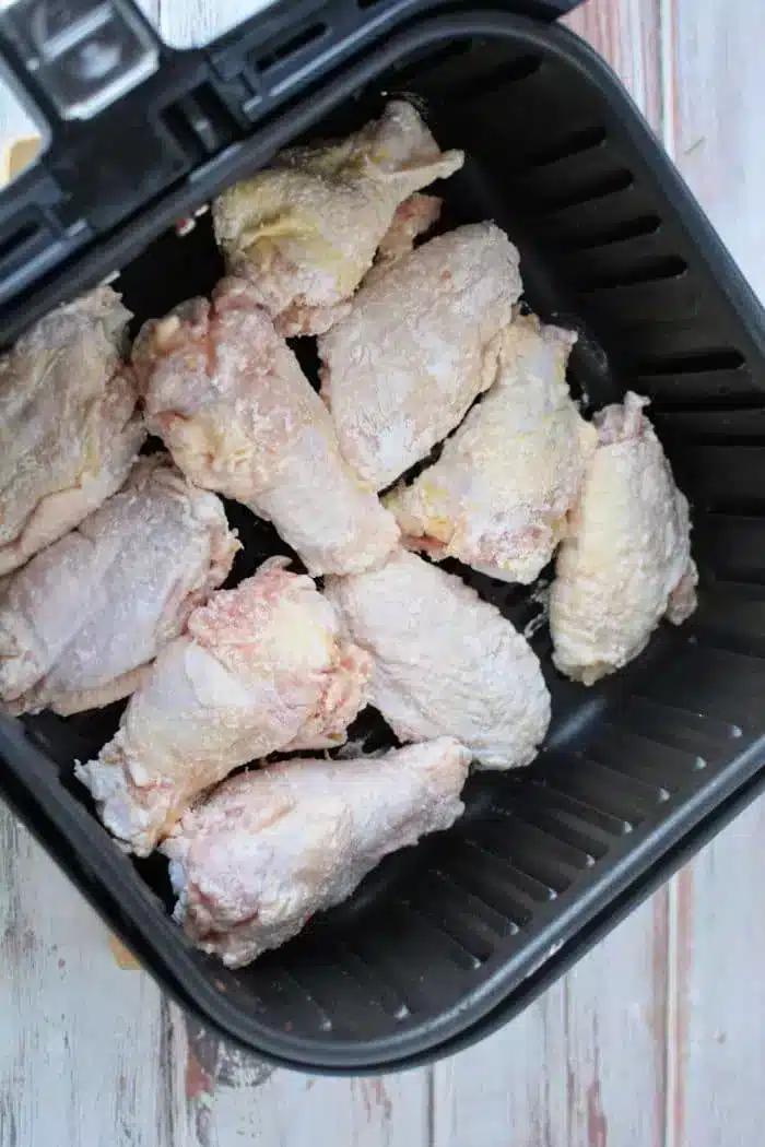 A close-up of raw, seasoned chicken wings dusted with baking powder, placed inside a black air fryer basket and ready for cooking. The countertop beneath reveals a light-colored surface with a faint wood grain pattern.