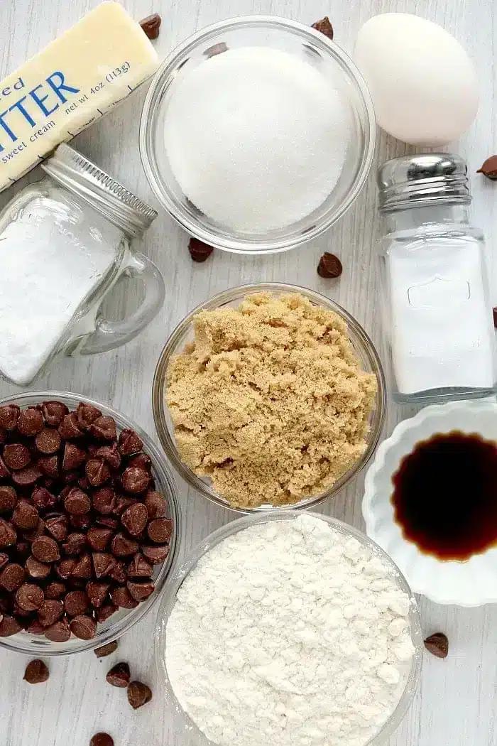 Ingredients for baking air fryer cookies are arranged on a surface: a stick of butter, a jar of sugar, brown sugar in a bowl, flour in another bowl, chocolate chips, an egg, a jar of baking soda, salt in a shaker, and a small bowl of vanilla extract.