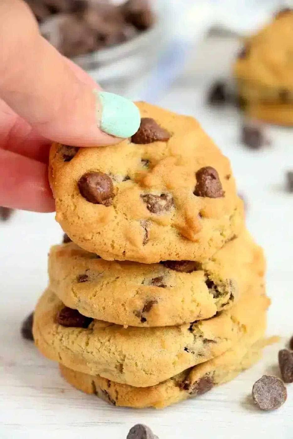 A hand with mint-green nail polish holds a chocolate chip cookie above a stack of air fryer cookies.