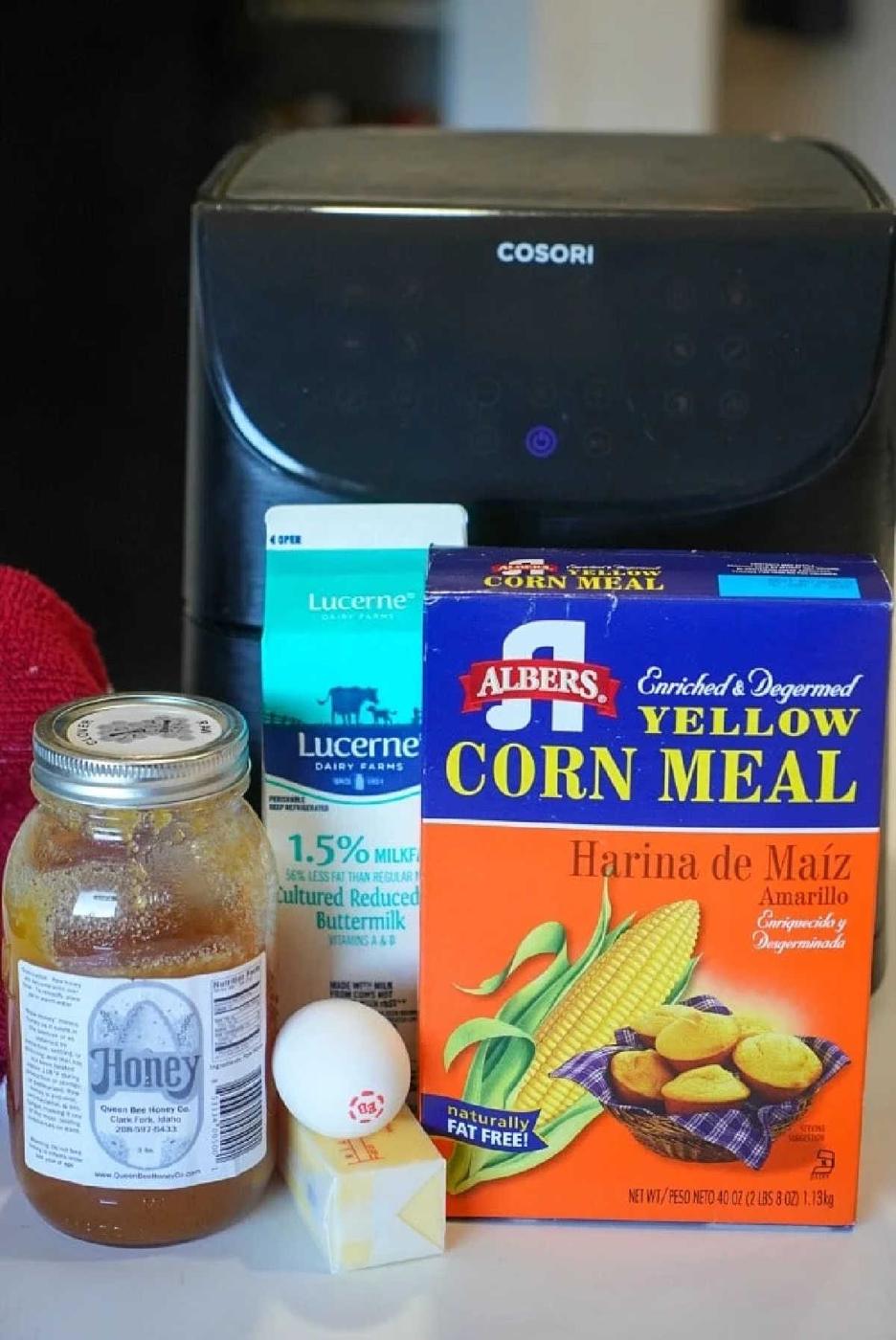 A carton of buttermilk, cornmeal box, honey jar, egg, and butter stick are arranged in front of an air fryer—perfect ingredients for making delicious air fryer cornbread.