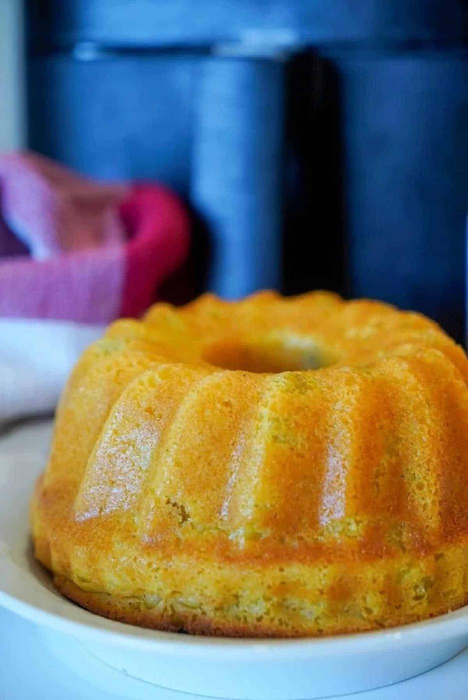 A golden brown air fryer cornbread bundt cake sits on a white plate, with a towel and dark background behind it.