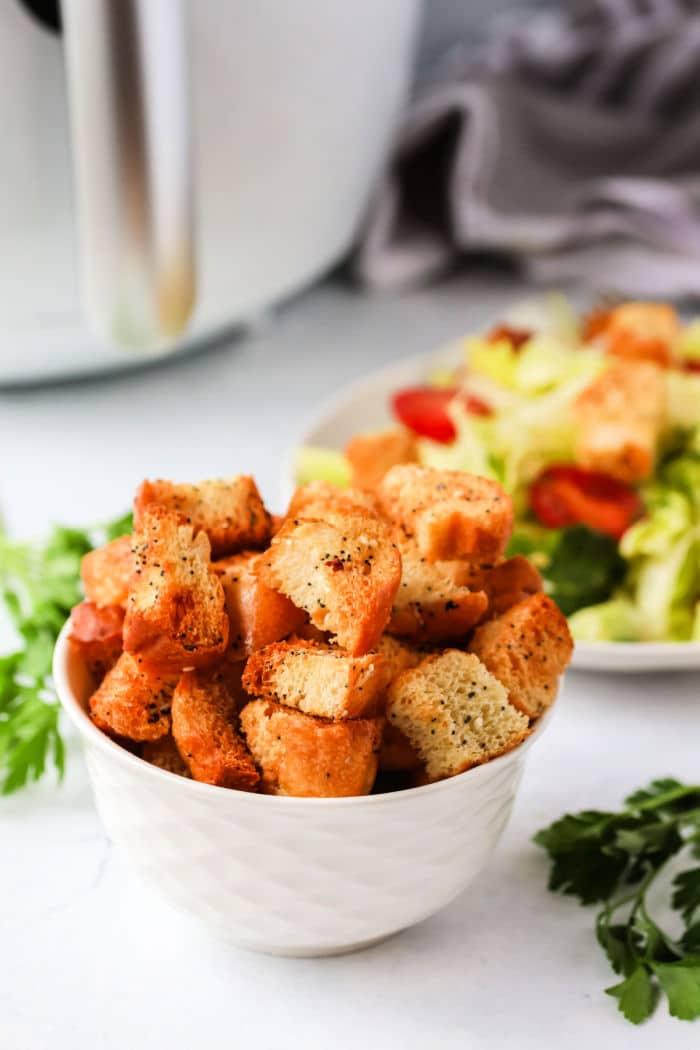 A bowl of perfectly seasoned air fryer croutons sits beside a salad overflowing with fresh tomatoes and crisp lettuce, all resting on a pristine white countertop embellished with scattered herbs.