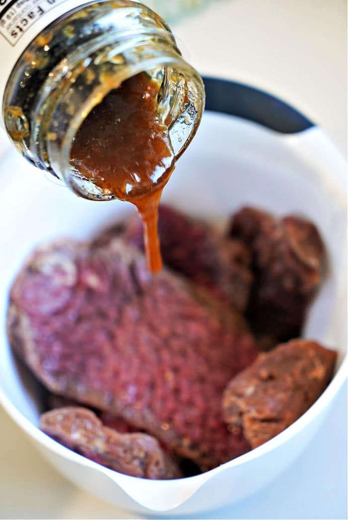 teriyaki sauce is being generously poured over raw cube steak in a white bowl, getting it prepped for the air fryer.