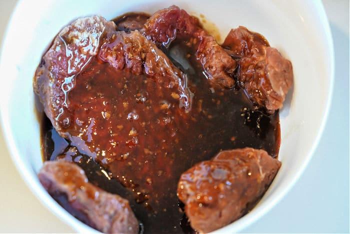 Raw cube steak pieces marinating in a dark, thick sauce in a white bowl, perfect for an air fryer feast.