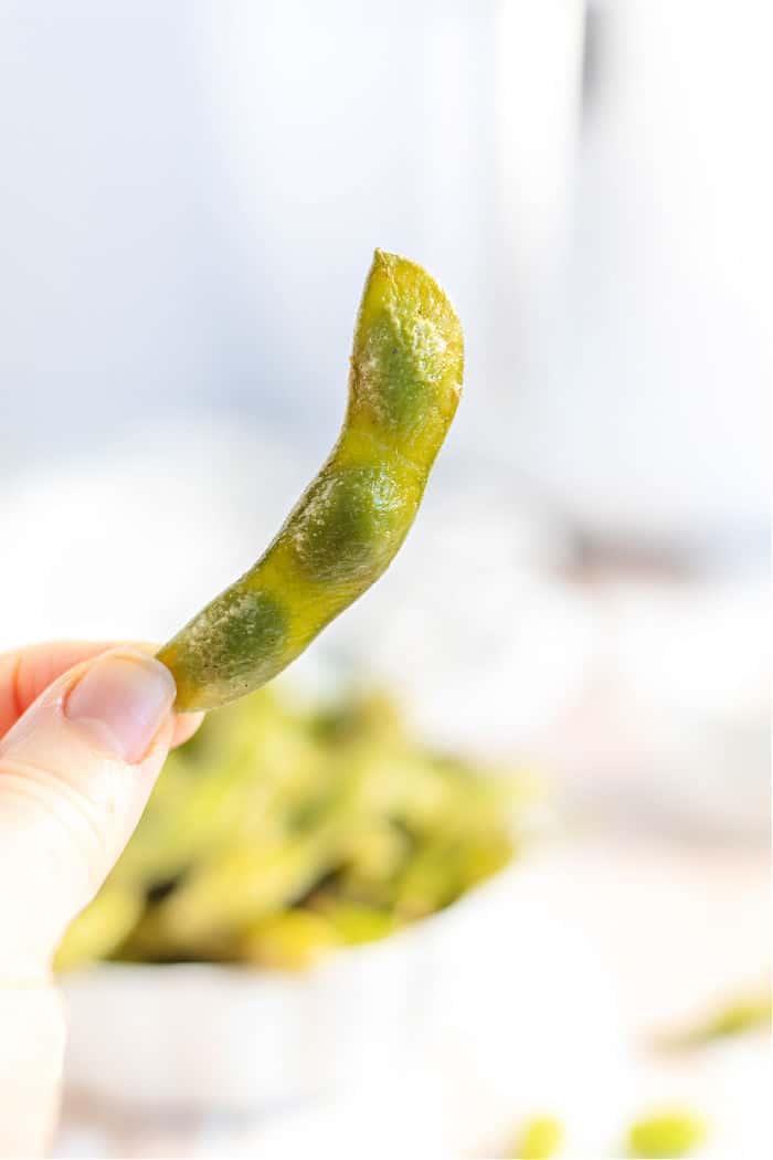 A hand holds a pod of fresh green edamame, perfect for preparing air fryer edamame, against a softly blurred background.