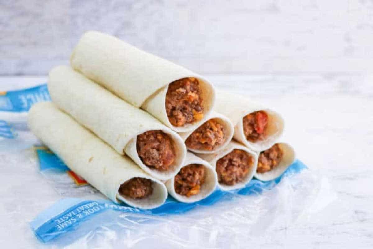 Eight air fryer flautas, filled with beef and cheese, are stacked on a blue plastic wrapper against a light background.