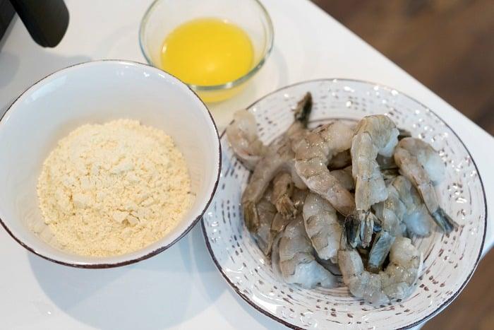 A bowl of raw shrimp awaits its transformation next to a bowl of flour and a small bowl of melted butter on a white countertop, ready for the air fryer to create perfectly fried shrimp.