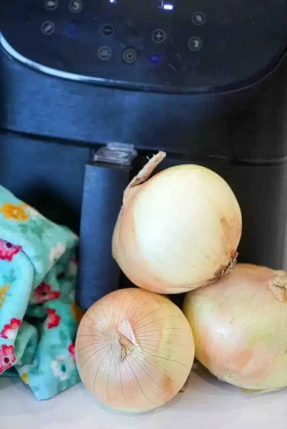 Three yellow onions sit in front of a black air fryer, perfect for making crispy air fryer onions, with a colorful cloth adding a pop of color to the left.