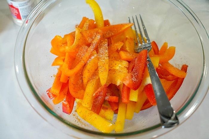 Sliced red, yellow, and orange air fryer peppers in a glass bowl with pepper seasoning and a fork.
