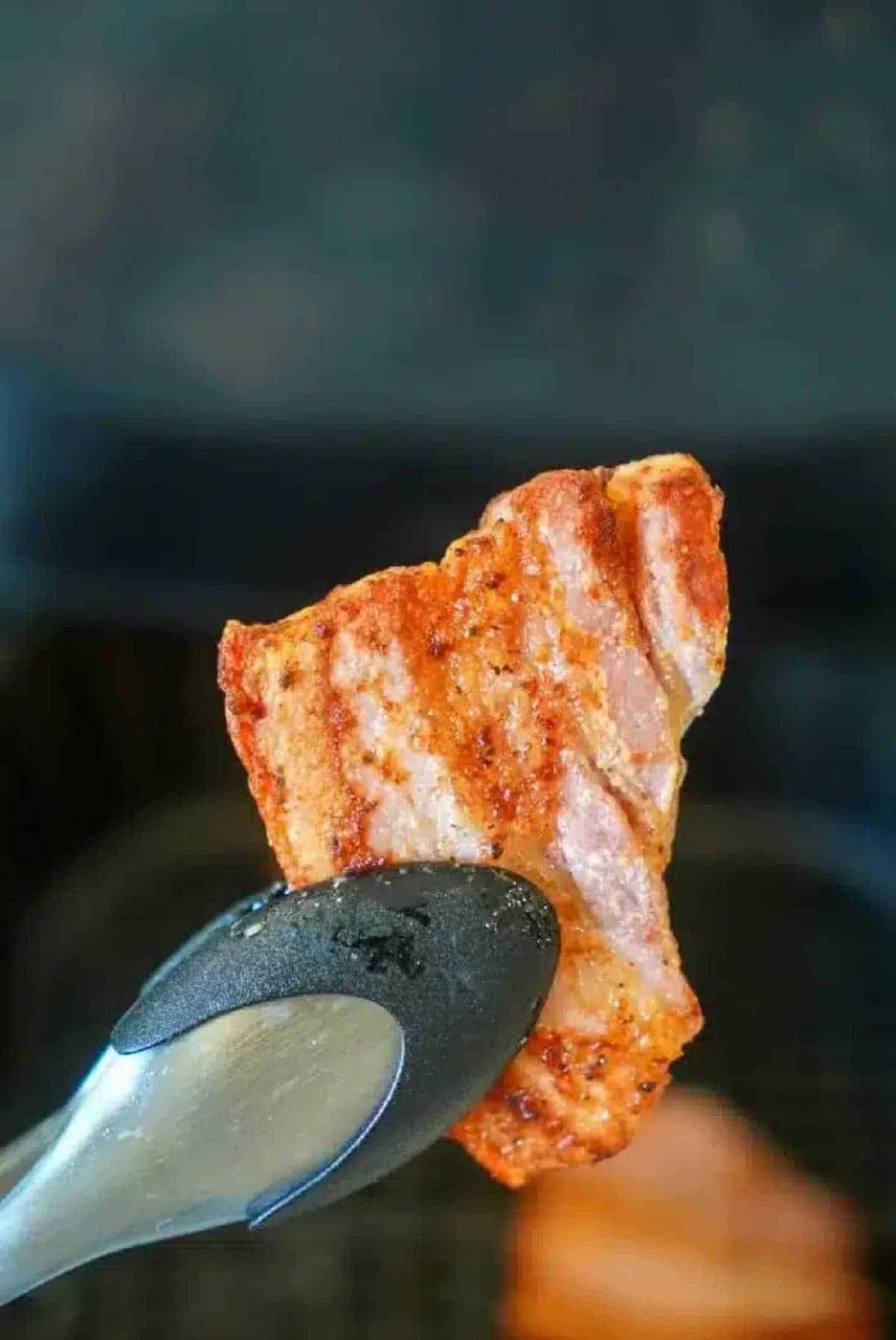 Metal tongs holding a grilled, seasoned piece of air fryer pork belly with crisp grill marks against a blurred background.
