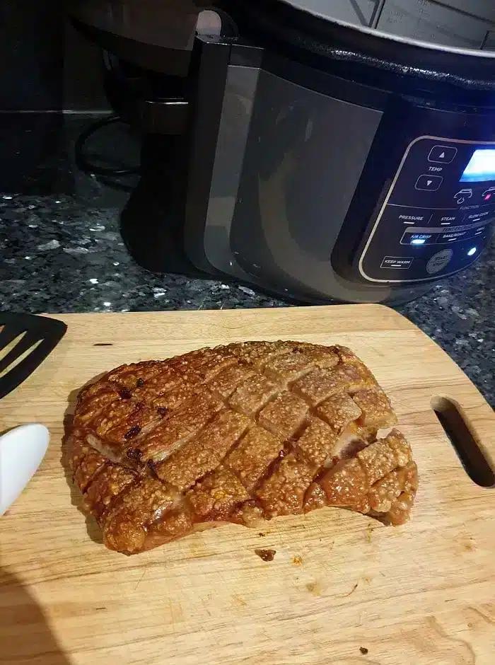 A wooden cutting board holds a piece of cooked pork belly with crispy, cross-hatched crackling. A spatula handle is visible on the left, and a slow cooker with digital controls sits in the background on a dark kitchen countertop, highlighting the results of perfectly air-fried pork belly.