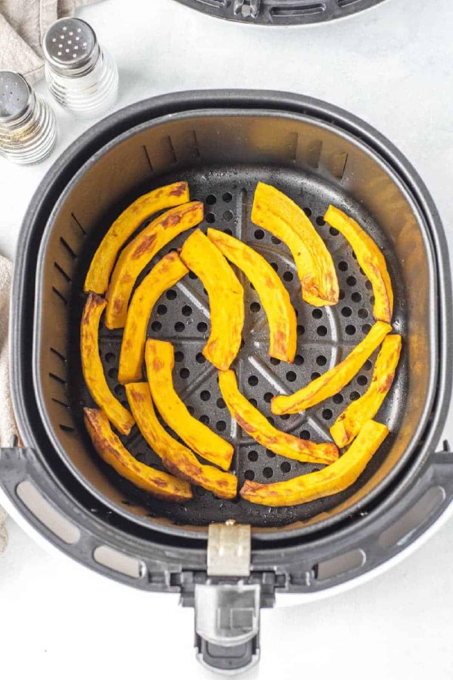 Air fryer pumpkin slices are neatly arranged in the basket, ready to roast, with salt and pepper shakers nearby for seasoning.
