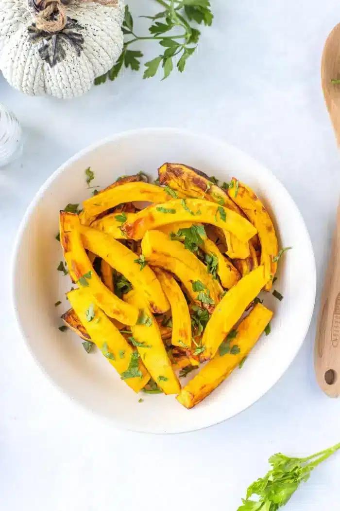 A white bowl contains roasted yellow squash garnished with chopped green herbs on a white surface. There is a wooden spoon on the right, and part of a decorative pumpkin and parsley can be seen in the top left corner, hinting at air fryer pumpkin slices to complete the autumnal scene.