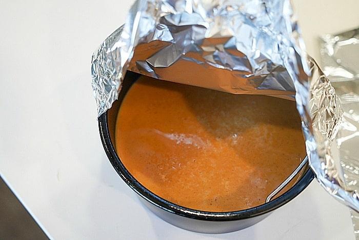A bowl of crustless pumpkin pie, is partially covered by aluminum foil on a white surface.