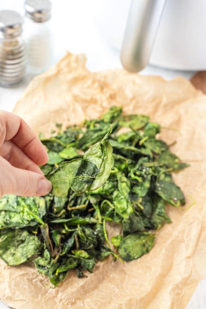 A hand holds a crisp spinach leaf over a pile of perfectly air-fried spinach on parchment paper, showcasing the delicious result of the air fryer spinach method.