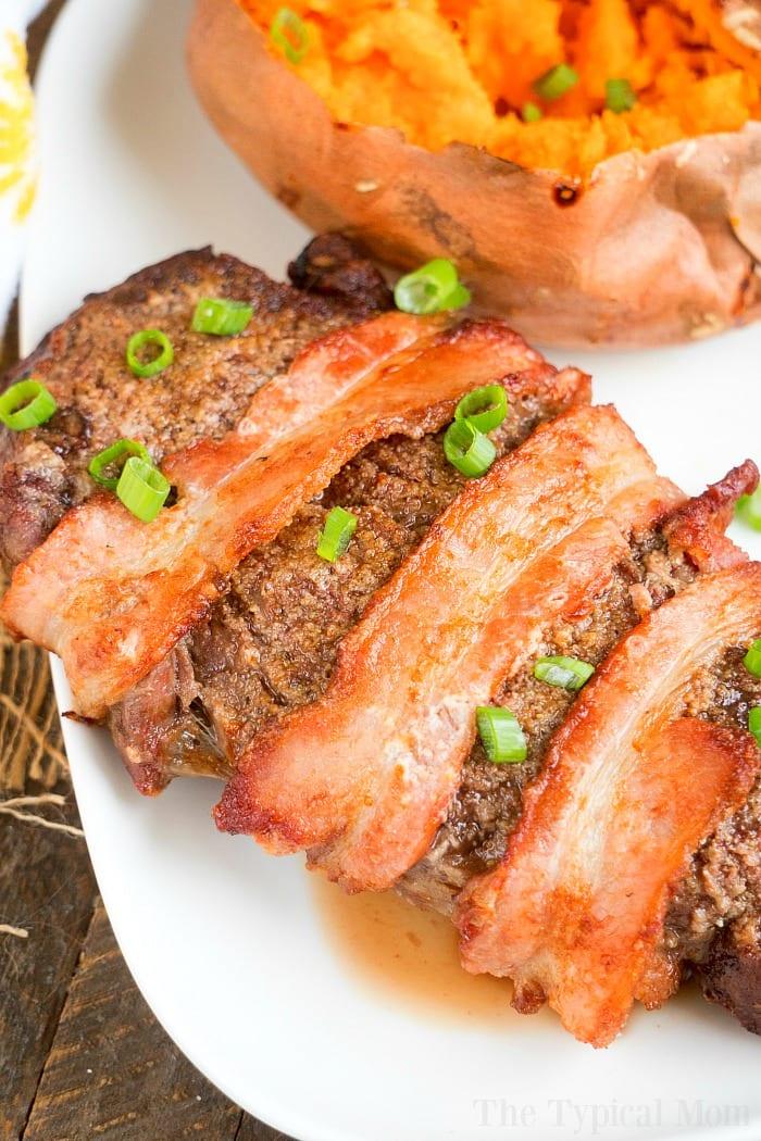 Bacon-wrapped steak garnished with green onions sits elegantly beside a baked sweet potato on a white plate, reminiscent of an air fryer steak in its perfection.
