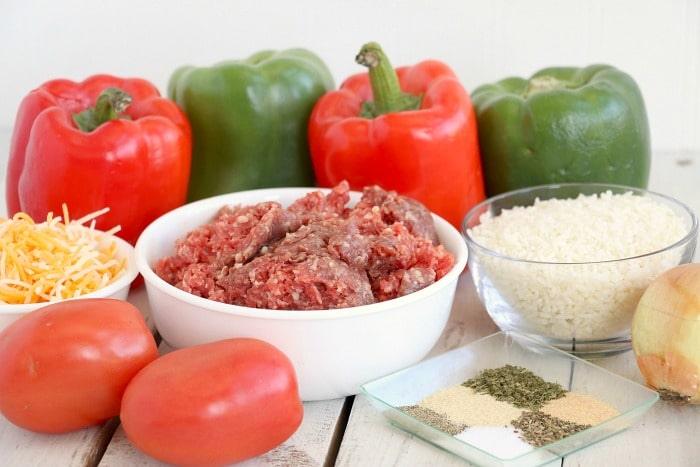 Bell peppers, ground meat, shredded cheese, tomatoes, uncooked rice, spices, and an onion on a white surface—perfect ingredients for delicious air fryer stuffed peppers.