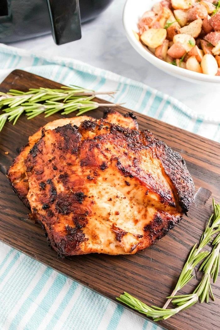 air fried boneless turkey breast with a charred exterior rests on a wooden board, garnished with sprigs of rosemary. A white bowl filled with roasted potatoes and herbs sits in the background, placed on a white and blue striped cloth.