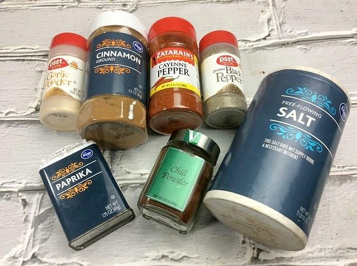 A collection of spice containers on a white brick surface, perfect for an air fryer wings recipe, includes garlic powder, ground cinnamon, cayenne pepper, black pepper, paprika, chili powder, and salt. Each container showcases distinct branding and labels.