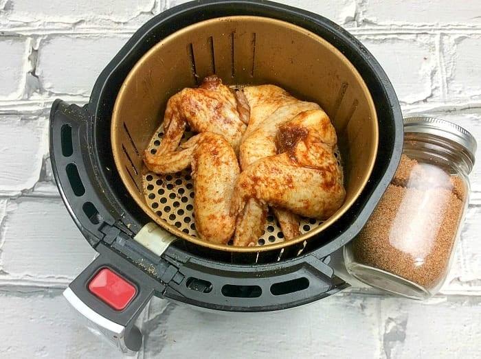 An air fryer wings recipe comes to life as a basket of perfectly seasoned chicken wings sits on a white brick background, accompanied by a jar filled with flavorful seasoning mix.