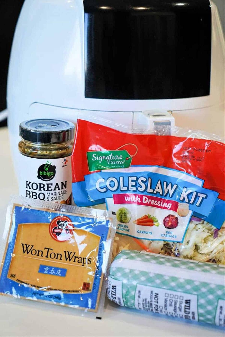 A jar of Korean BBQ sauce, coleslaw kit, wonton wraps, and ground pork in front of an air fryer—perfect for making flavorful air fryer won tons at home.