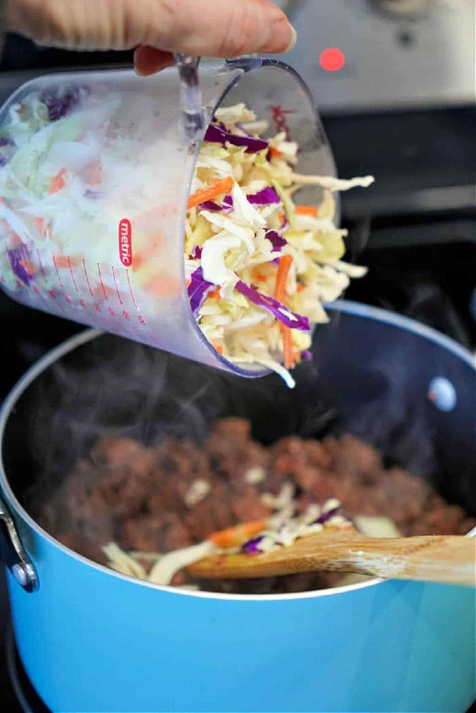 Hand pouring shredded cabbage mix from a measuring cup into a blue pot with cooked ground meat, perfect for a savory filling in homemade air fryer won tons.