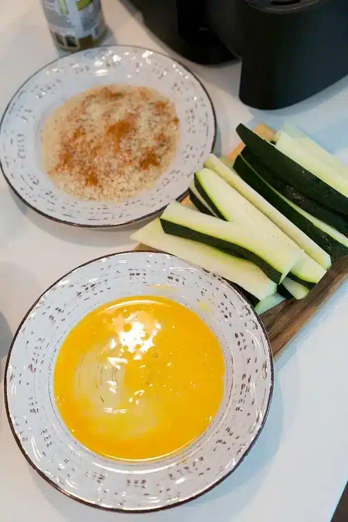 Two bowls on a white surface, one with beaten eggs, the other with a breadcrumb mixture. Next to the bowls are sliced zucchini on a cutting board, ready to become delicious air fryer zucchini fries.