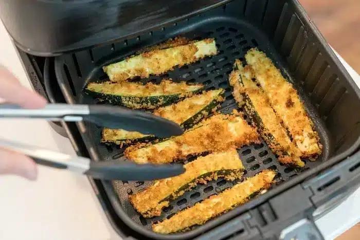 A person uses tongs to remove crispy air fryer zucchini fries from the basket. The zucchini sticks are coated in a golden-brown breading and evenly spaced in the black basket, with the air fryer lid open to reveal the delicious contents.