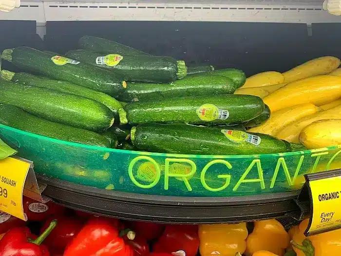 A grocery store display features green zucchini with produce stickers and yellow squash, perfect for making air fryer zucchini fries. A partially visible yellow sign reads u0022ORGANIC.u0022 Red bell peppers are visible in the bottom of the frame.
