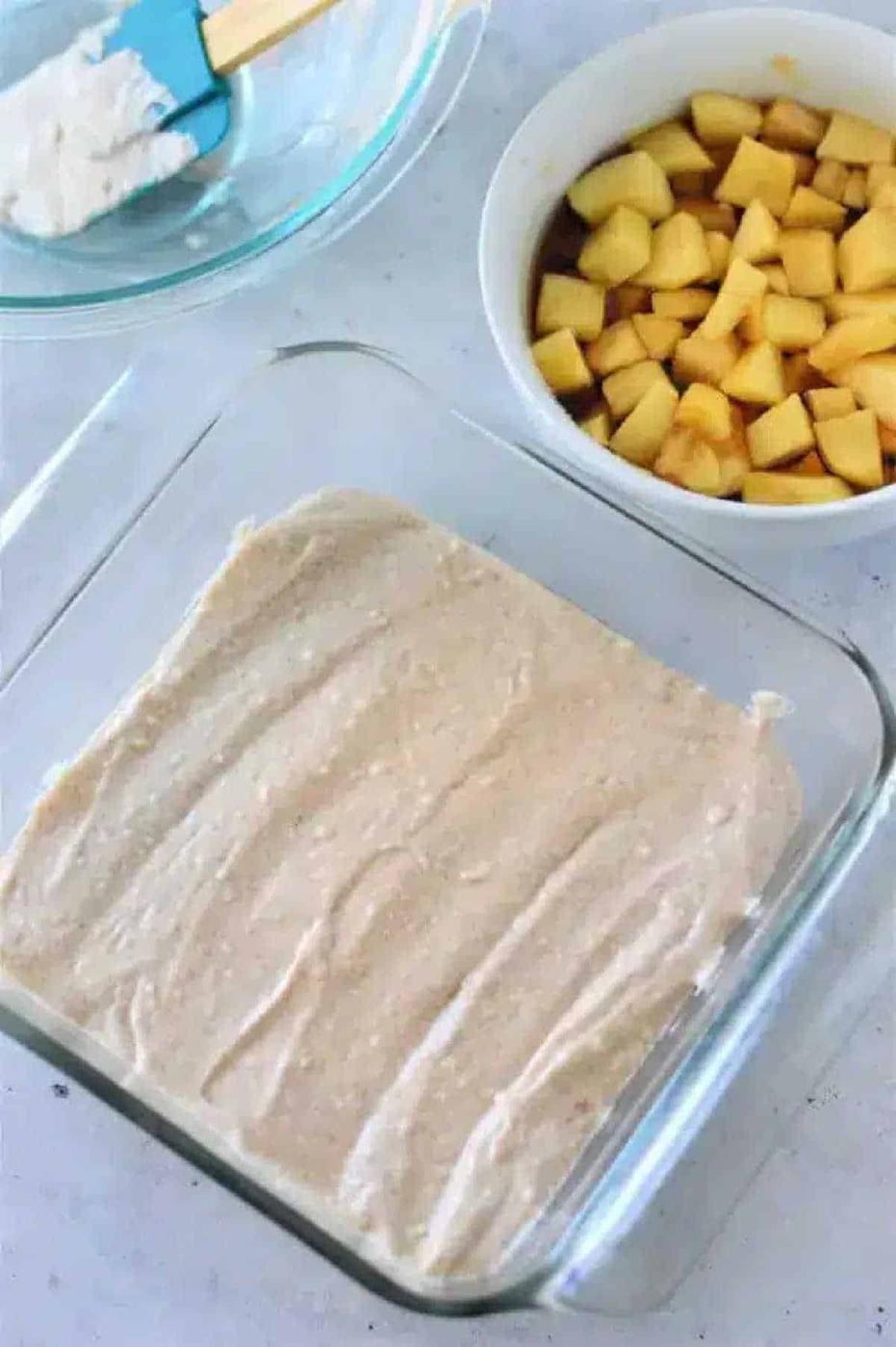 Glass baking dish with Apple Cobbler with Bisquick batter, bowl of chopped apples, and mixing bowl with spatula on a white surface.