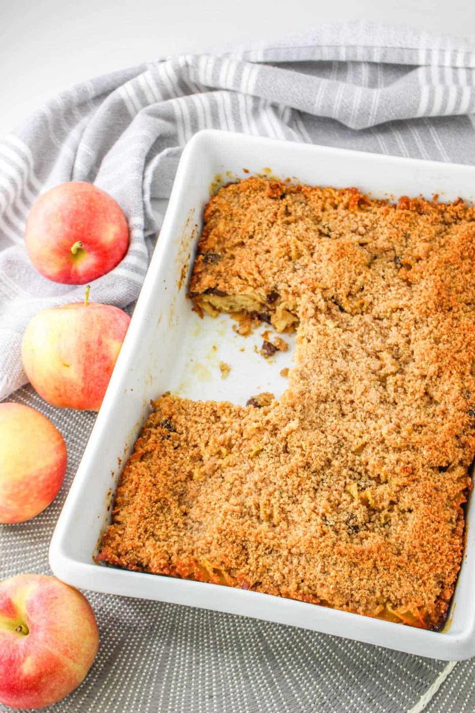 A delightful apple kugel sits invitingly in a white baking dish, reminiscent of a comforting apple kugel, with three fresh apples and a charming striped cloth nearby.