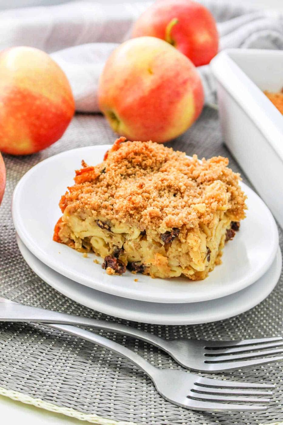 A slice of apple kugel cake, reminiscent of a classic apple kugel, sits on a white plate surrounded by whole apples and forks.