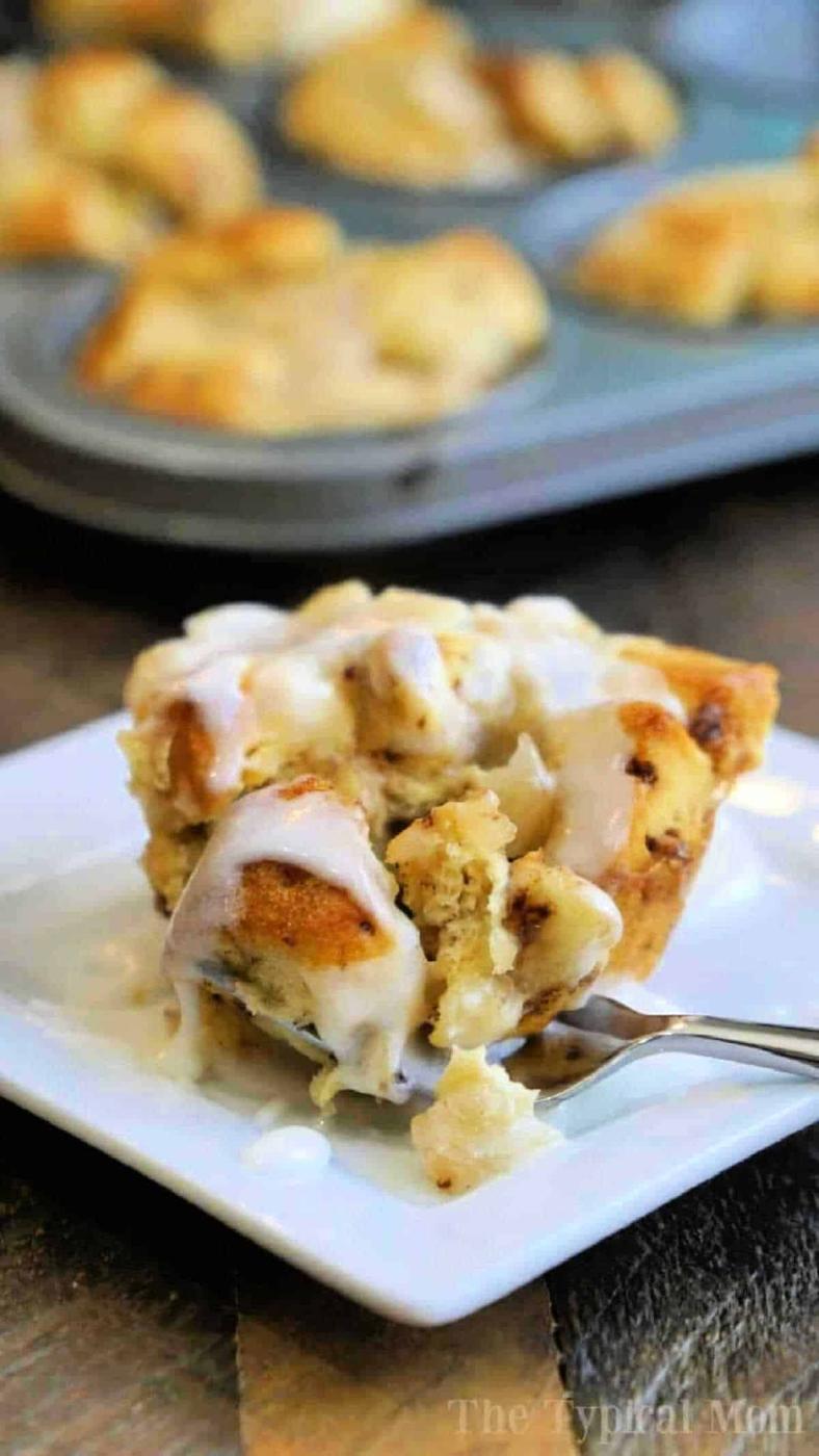 A gooey cinnamon roll muffin with icing on a white plate, with more monkey bread muffins in a tray in the background.