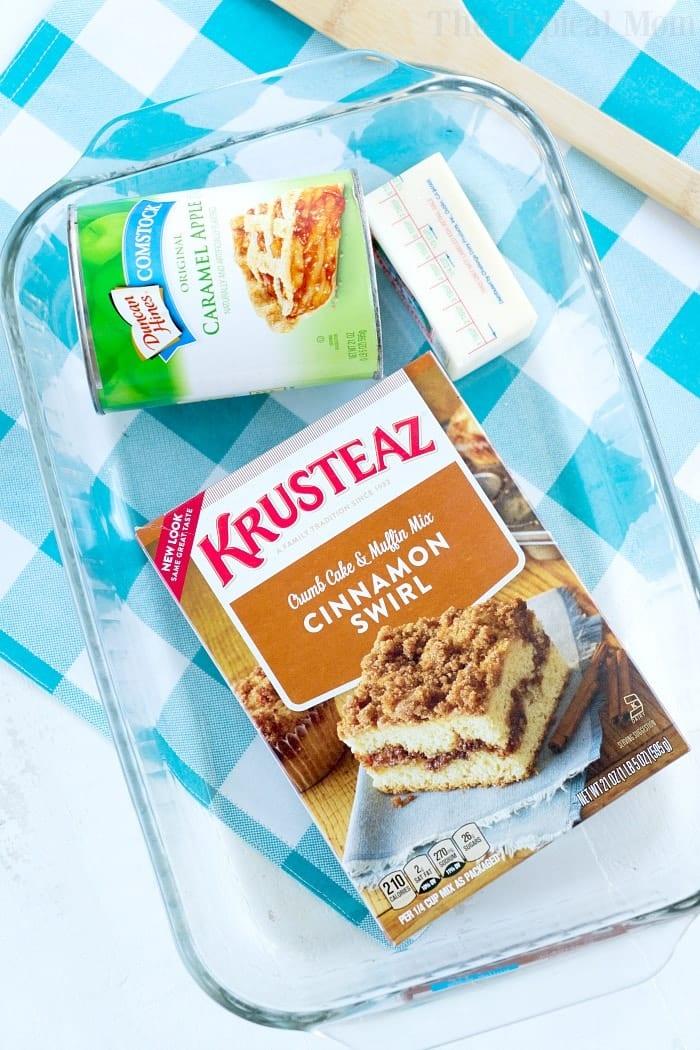 Box of Krusteaz cinnamon swirl mix, caramel apple pie filling, and butter in a glass baking dish on a blue checkered cloth—perfect for making an easy apple spice dump cake.