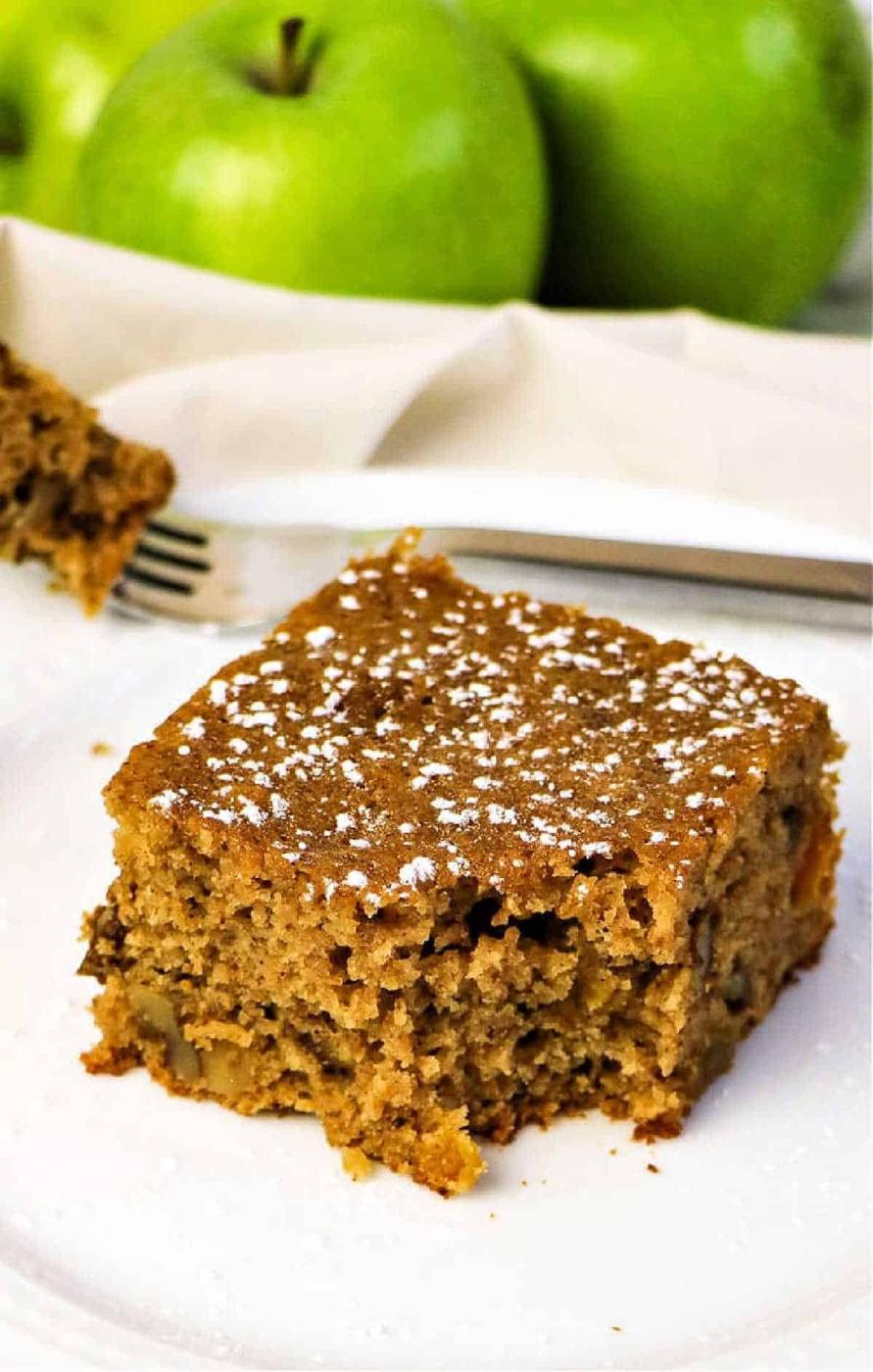 A slice of applesauce spice cake dusted with powdered sugar sits on a white plate, with green apples in the background.