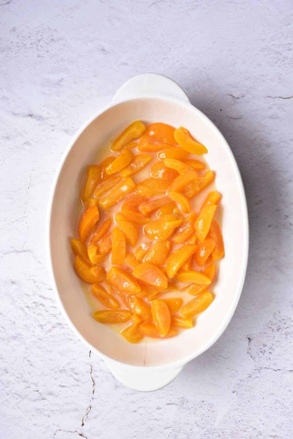 Oval dish with sliced apricots in syrup, reminiscent of an apricot cobbler, placed on a textured white surface.