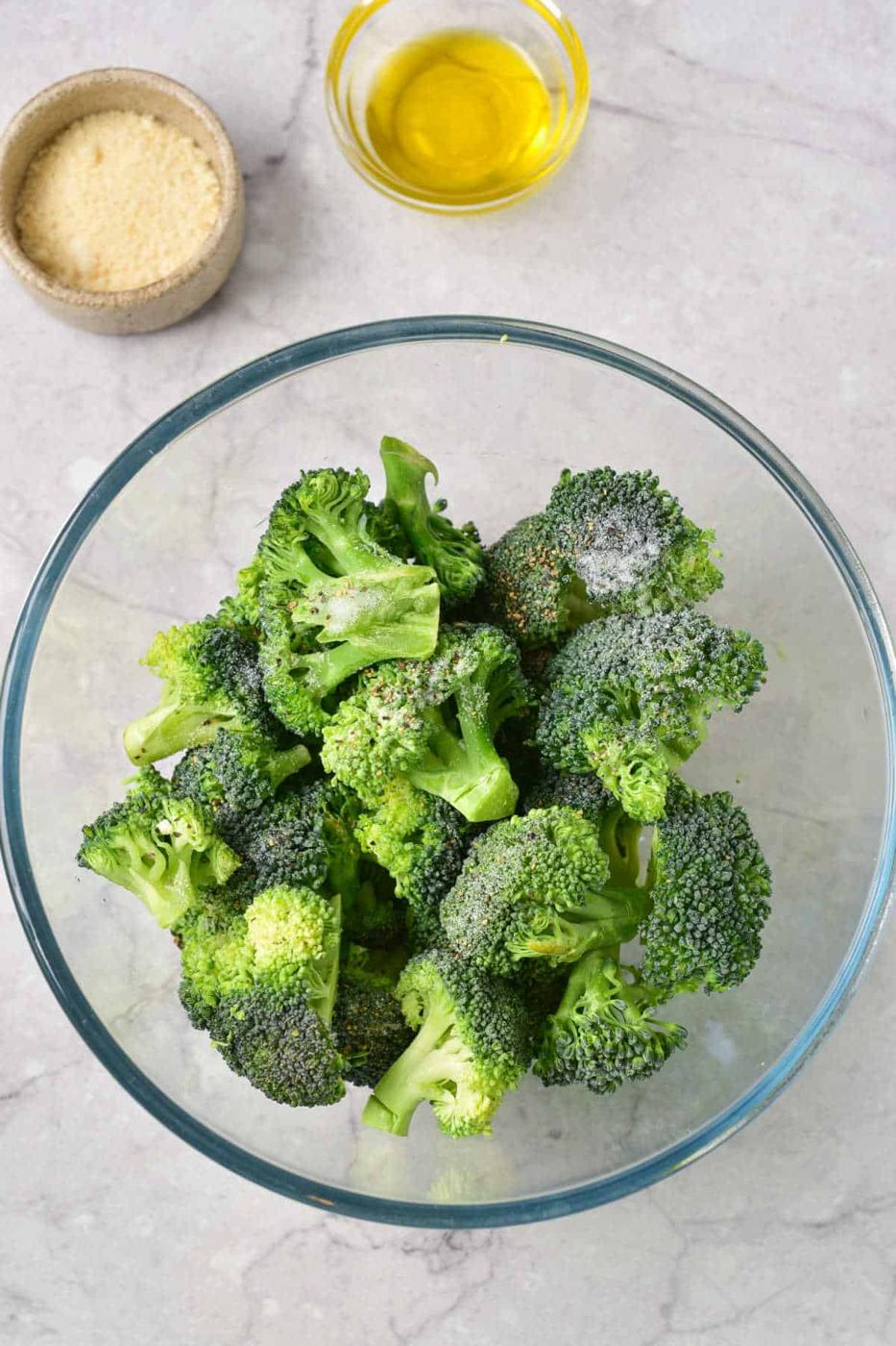 A glass bowl of raw broccoli florets with seasonings, perfect to bake frozen broccoli, sits next to bowls of olive oil and grated cheese.